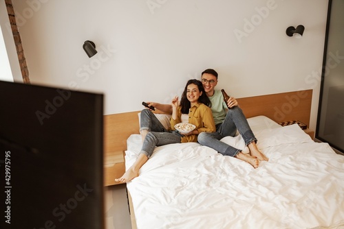 Lovely couple with plate of pop conr while watching movie on tv, sitting on bed. Happy young family moved to new apartment photo