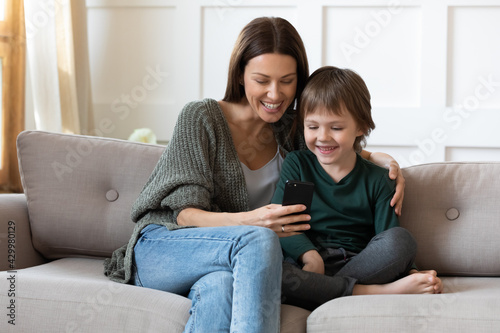 Happy young mom and small 7s son relax on sofa in living room have fun use browse cellphone together. Smiling Caucasian mother and little boy child have webcam digital video call on smartphone.