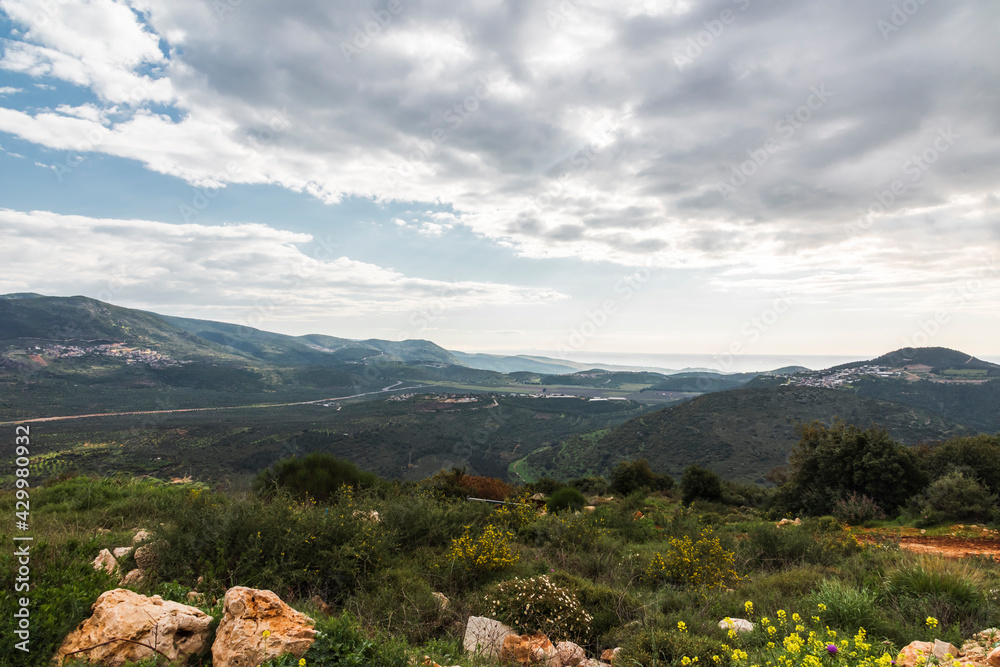 Obraz premium A view of a mountain range and a green valley in the morning at sunrise, against a dramatic backdrop of blue skies and clouds. North District Israel. High quality photo. Travel concept hiking