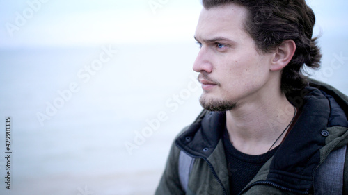 Attractive blue-eyed young man with beard and curly hair lone portrait on a windy cliff above dark sea. Looking worried, He look up on troubled water and cloudy sky with a hope at late autumn
