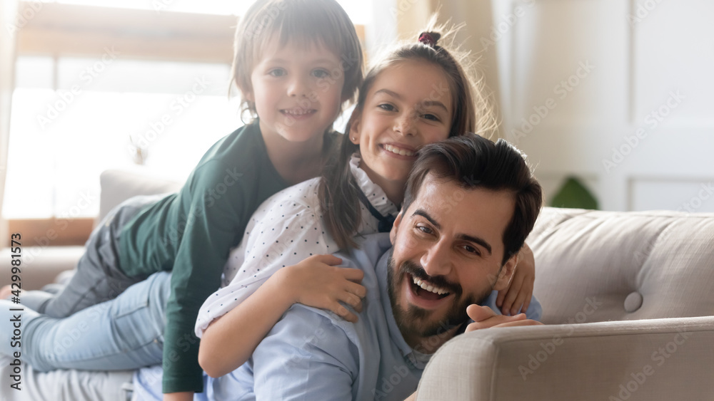 Portrait of smiling young Caucasian single dad and two small kids lying on sofa have fun feeling playful. Happy loving father and little children relax rest on couch at home on family weekend. Portrait of smiling young Caucasian single dad and two small kids lying on sofa have fun feeling playful. Happy loving father and little children relax rest on couch at home on family weekend.