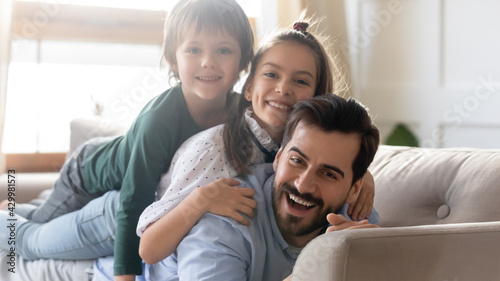 Portrait of smiling young Caucasian single dad and two small kids lying on sofa have fun feeling playful. Happy loving father and little children relax rest on couch at home on family weekend.
