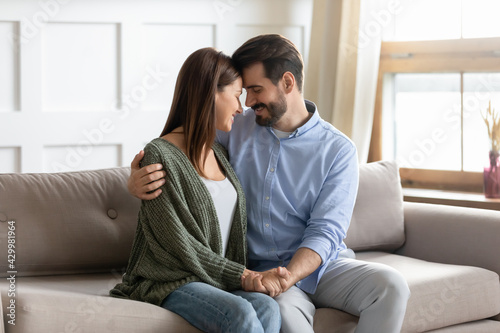 Happy young couple sit relax on sofa at home hug and cuddle show love and care in relationships. Smiling millennial Caucasian man and woman rest on couch enjoy romantic close date. Family concept.