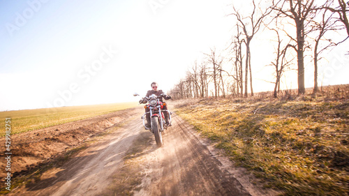 Young curly biker with red glasses drive ride on red chopper through the desert dirt road in late autumn sunset light