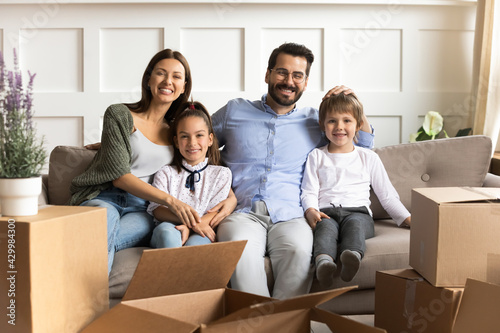 Portrait of smiling young parents with two little children relax in living room moving to new house or apartment. Happy Caucasian family with small kids relocate to own home. Relocation concept.