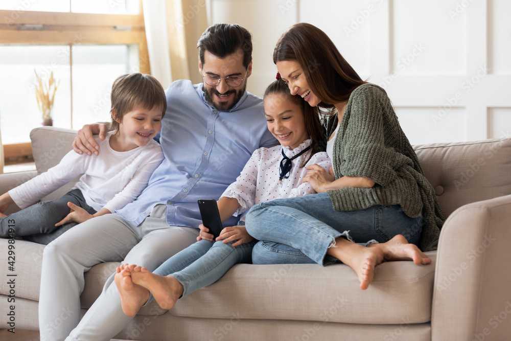 Smiling young Caucasian family with two children relax on couch use cellphone talk speak on video call together. Happy parents with kids browse smartphone have webcam call. Technology concept. Smiling young Caucasian family with two children relax on couch use cellphone talk speak on video call together. Happy parents with kids browse smartphone have webcam call. Technology concept.