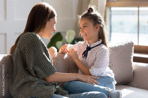 Happy young Caucasian mother and teen daughter sit relax on sofa talking sharing secrets. Smiling mom or nanny and teenage girl child rest on couch speak enjoy family weekend bonding at home.