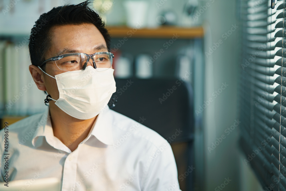 Medium close up portrait of Asian man wearing eye protection and protective face mask looking through out the windows with worried while sitting in the room. Hope and despair covid-19 pandemic. Medium close up portrait of Asian man wearing eye protection and protective face mask looking through out the windows with worried while sitting in the room. Hope and despair covid-19 pandemic.