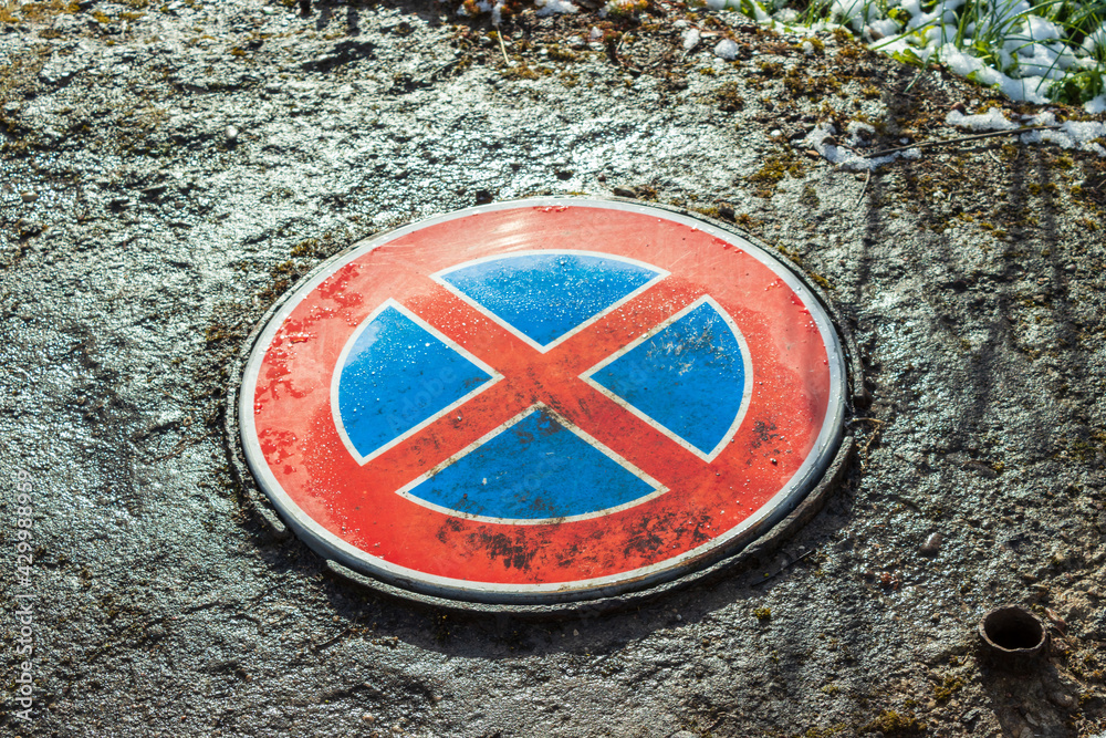 Wet shining red road sign as sewerage cover, red cross over blue metal circle