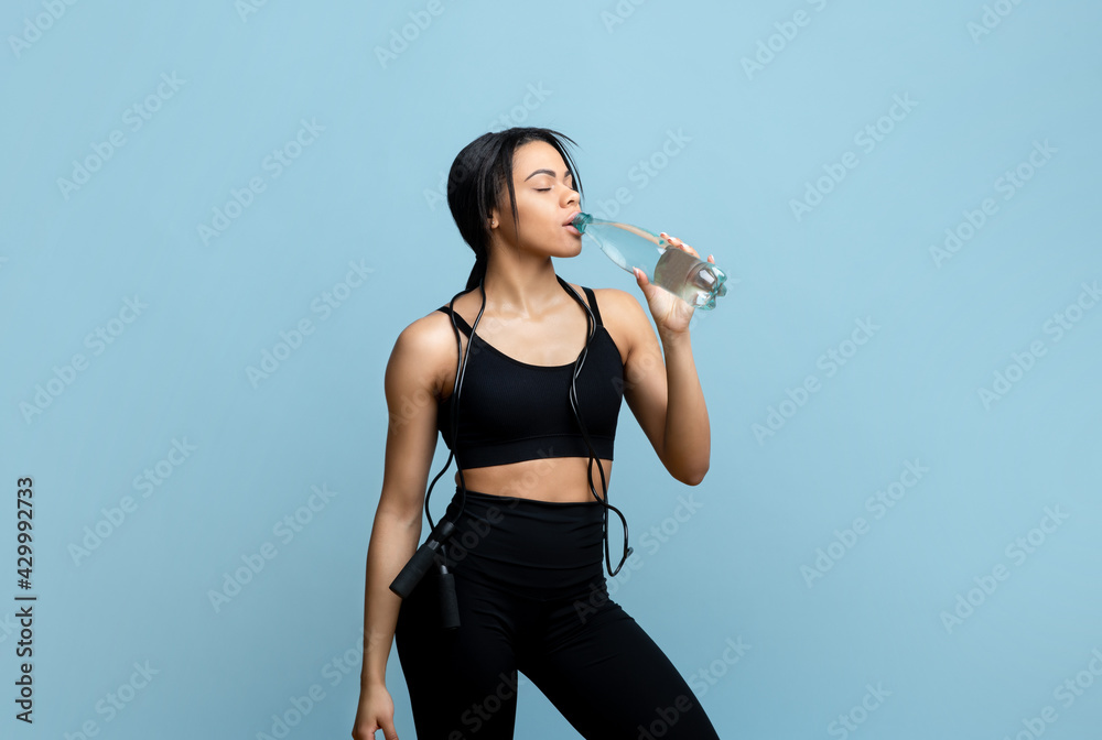 Sports hydration concept. Sporty african american woman drinking mineral water from bottle at blue studio background Sports hydration concept. Sporty african american woman drinking mineral water from bottle at blue studio background
