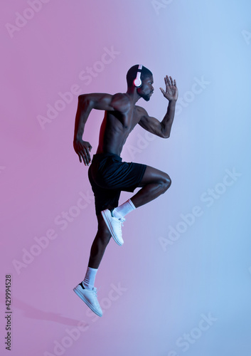 Full length of athletic black sportsman in headphones running and listening to music in neon lighting, side view