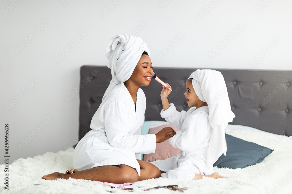 Full length portrait of African American mother and daughter in bath robes and towels applying makeup on bed at home Full length portrait of African American mother and daughter in bath robes and towels applying makeup on bed at home