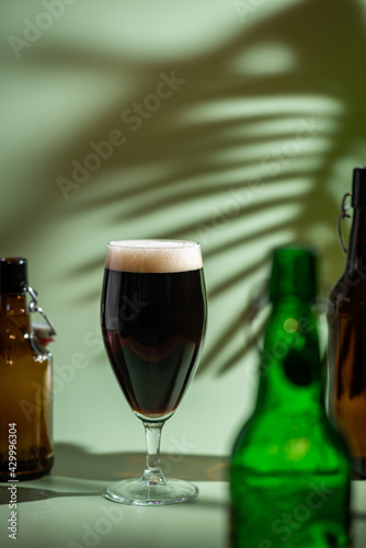 Glass of dark beer with foam head and empty bottles on colorful green and orange background  bright shadows
