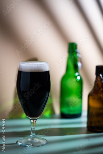 Glass of dark beer with foam head and empty bottles on colorful green and orange background, bright shadows