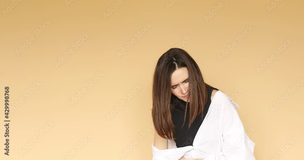Painful cramp in stomach. Sick unhealthy girl clasping belly and suffering from severe abdominal pain, periods spasm, irritable bowel syndrome of diet. Indoor studio shot isolated on beige background