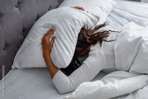 Stressed black woman covering head under pillow lying in bed photo