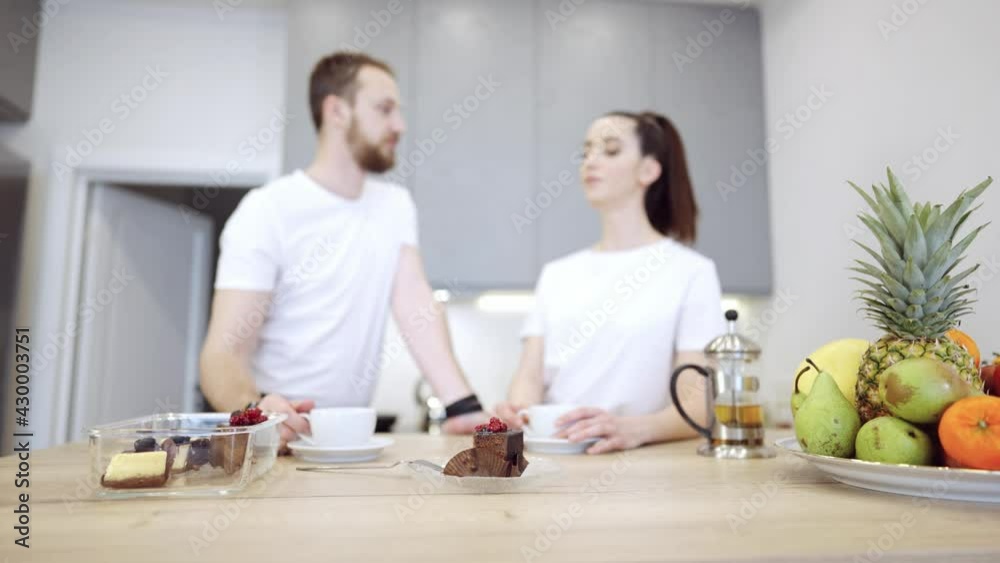Young cheerful couple in the kitchen drink tea and pouring tea into a cup from a teapot. There are fruits and vegetables on the wooden table. They discuss healthy food, vegan nutrition. Young cheerful couple in the kitchen drink tea and pouring tea into a cup from a teapot. There are fruits and vegetables on the wooden table. They discuss healthy food, vegan nutrition.