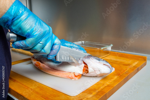 The hands of a male cook in hygienic gloves cut off the head of a salmon with a knife. Cutting fresh fish in the kitchen on a cutting board.