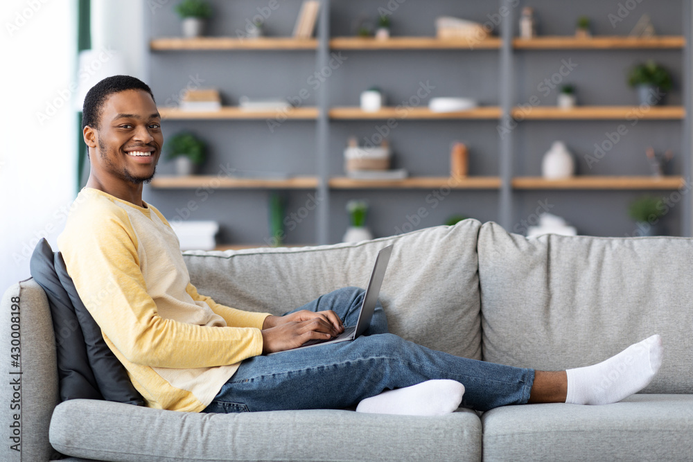 Fototapeta premium Side view of black guy reclining on couch with laptop