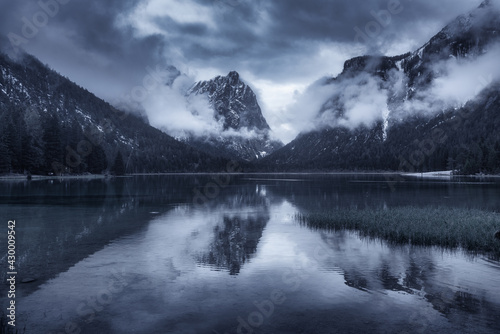 Mountain lake in overcast rainy day in spring. Dramatic landscape with lake, dark sky with low clouds, water with reflection, trees, high rocks in fog in summer. Moody scenery. Dolomites, Italy 