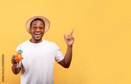 Amazing offer. Excited African American man with yummy summer cocktail pointing at blank space on yellow background
