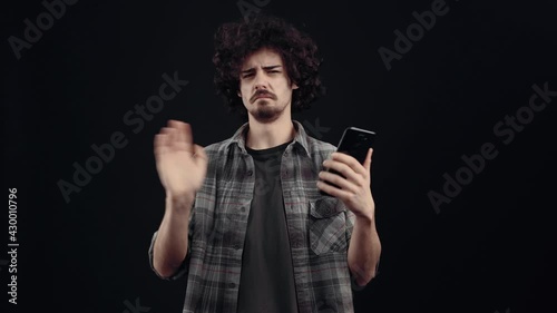 The irritated and frowning young man looks at the phone screen, receives a message that upsets him and he shakes his hand that no, no, no. Isolated on black background, Concept of life. People's