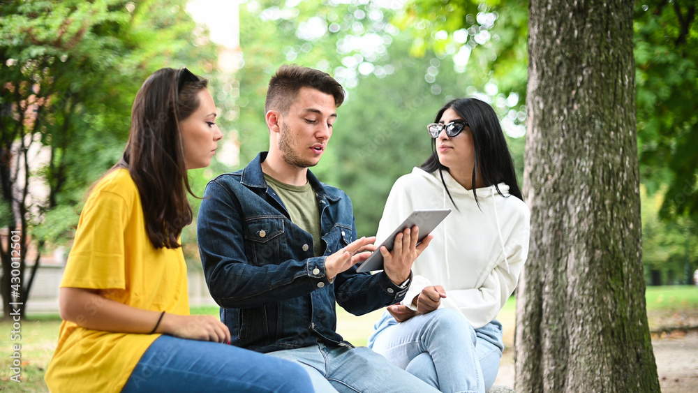 Three students studying together with a digital tablet sitting on a bench outdoor Three students studying together with a digital tablet sitting on a bench outdoor