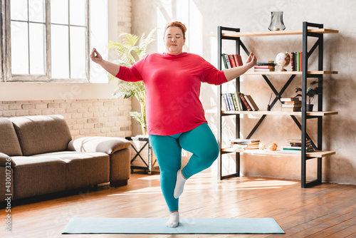 Obese young fat woman standing in yoga position at home. Chubby overweight young caucasian woman practicing fitness for losing weight and burning calories photo