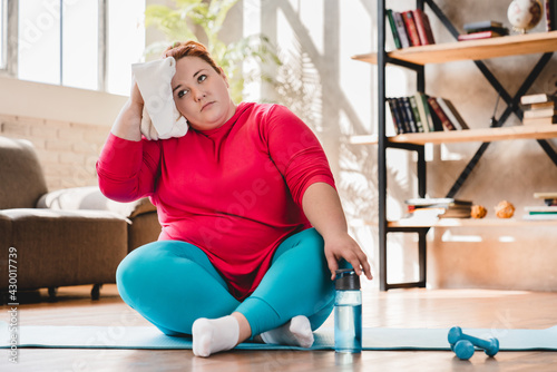Tired exhausted young caucasian fat woman sitting and relaxing after hard training at home for fat burning and losing weight. Chubby plump woman doing fitness exercises indoors