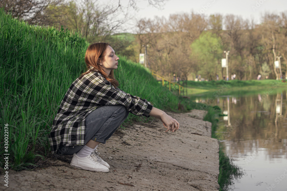 Portrait of a young girl on the background of the lake. The girl is sitting on the shore of the lake. Girl with red hair