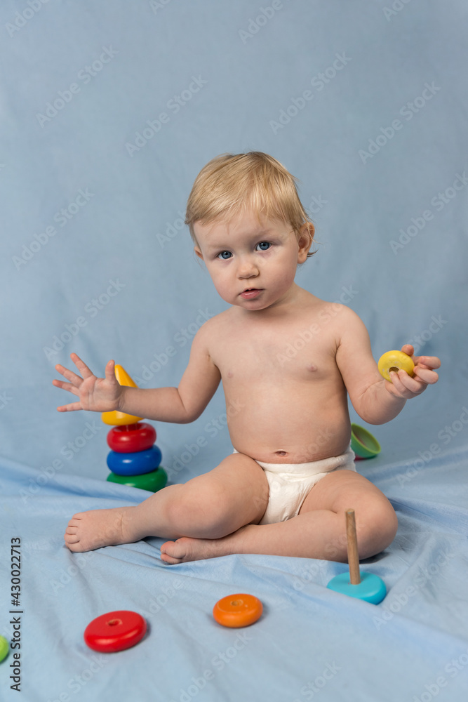 A little blonde girl in white diaper sits on a blue background and plays with a wooden pyramid made of colorful rings. Looking at the camera A little blonde girl in white diaper sits on a blue background and plays with a wooden pyramid made of colorful rings. Looking at the camera
