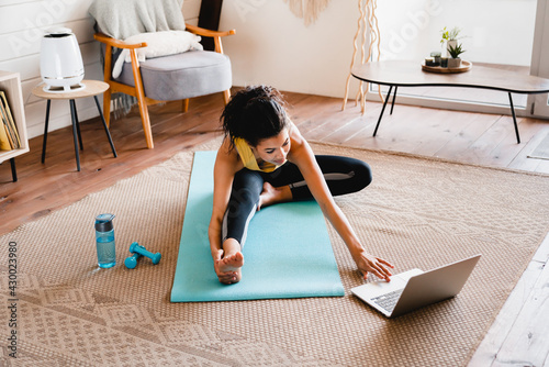 Young slim african girl stretching at home using laptop. Cheerful African woman stretching up In front of laptop, doing home workout, warming up for domestic fitness training, exercising  photo