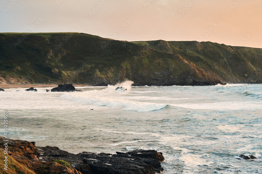 Landscape of a rolling sea at sunset with a lone surfer paddling up the waves.