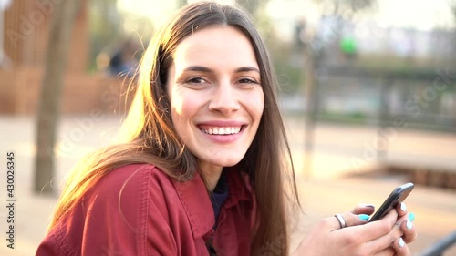 Portrait of a cheerful young woman texting on mobile phone and them turning and smiling in to the camera outdoors during the sunset 