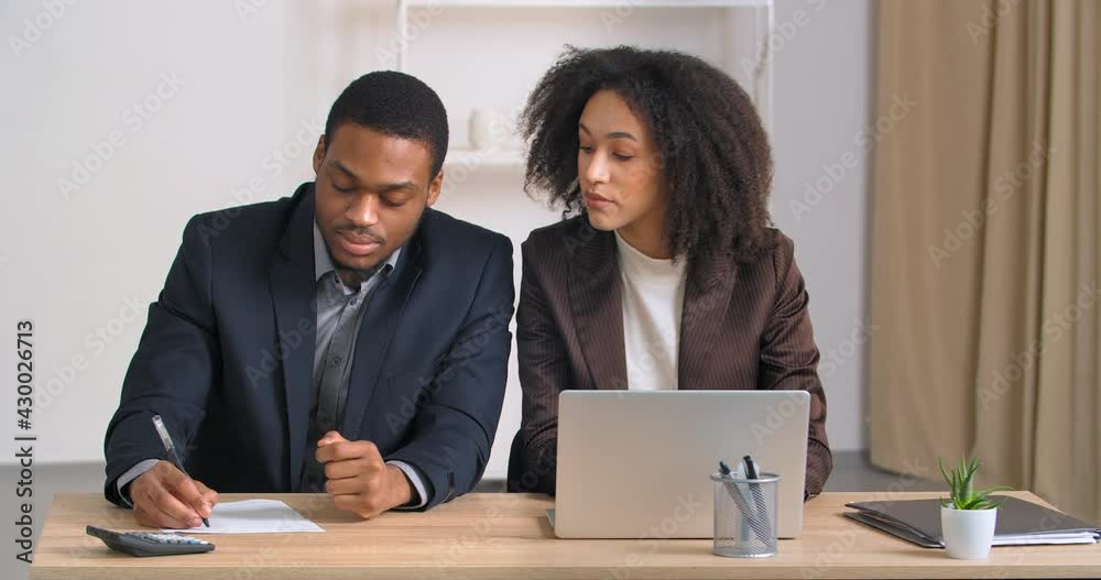 Afro american woman financial consultant accountant helps ethnic businessman cost accounting of expenses plan budget of company looks into laptop dictates numbers talking man writes report in document Afro american woman financial consultant accountant helps ethnic businessman cost accounting of expenses plan budget of company looks into laptop dictates numbers talking man writes report in document
