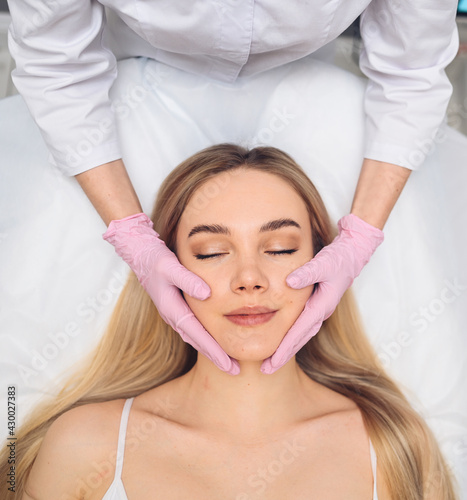 Closeup of beautician hands in gloves touching young woman's face. Plastic surgery concept. Facial beauty. Portrait of beautiful blonde female with perfect makeup and soft smooth skin. 