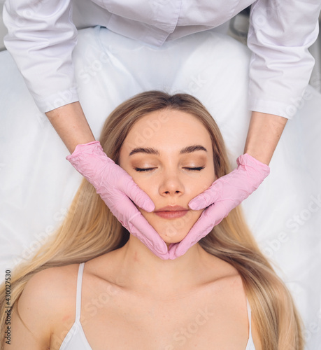Closeup of beautician hands in gloves touching young woman's face. Plastic surgery concept. Facial beauty. Portrait of beautiful blonde female with perfect makeup and soft smooth skin. 