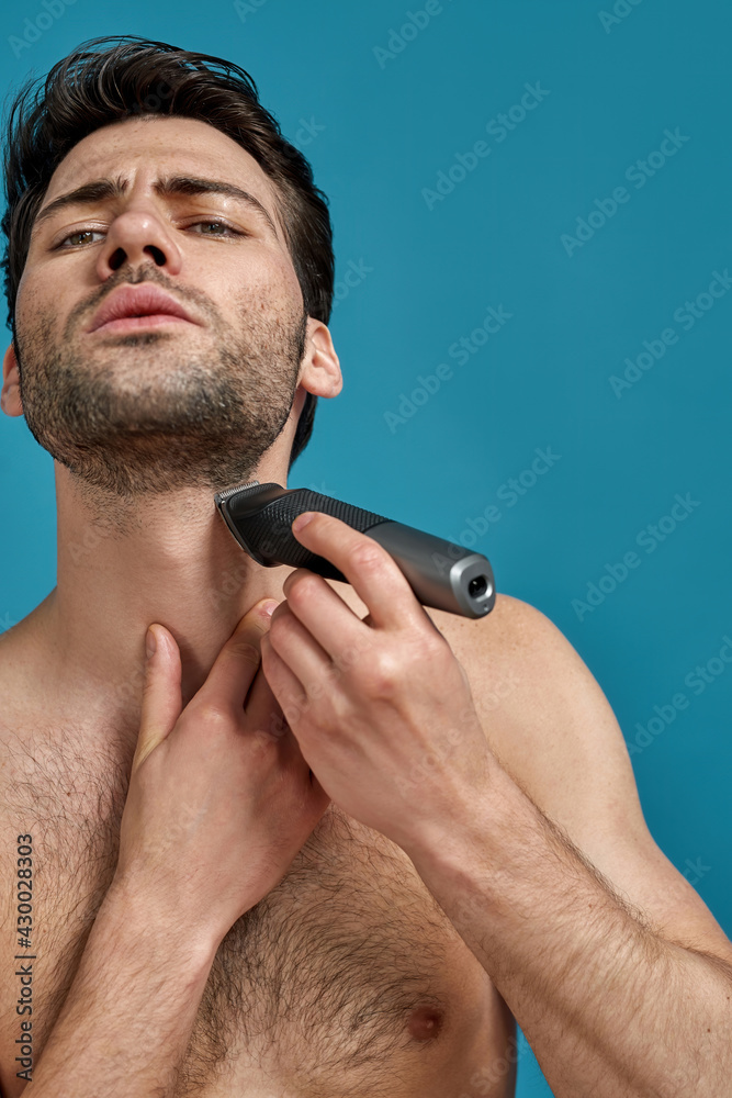Fototapeta premium Portrait of handsome naked man looking concentrated while using electric shaver isolated over blue background