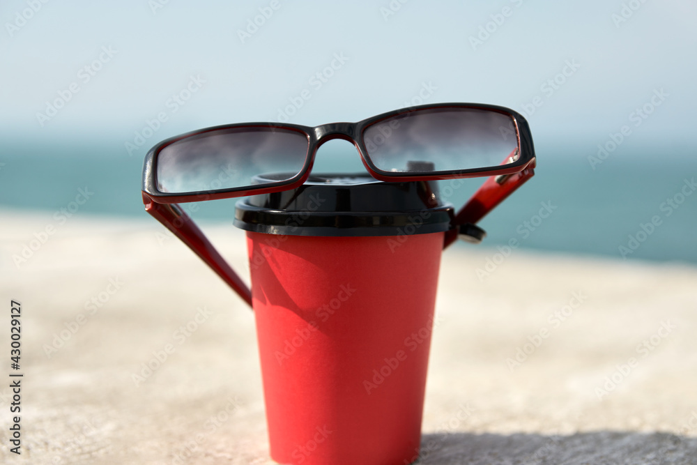  A disposable red coffee cup stands on the concrete parapet of the embankment. The cup is covered with sunglasses.In the background, the horizon line between the sea and the sky. Copy space.