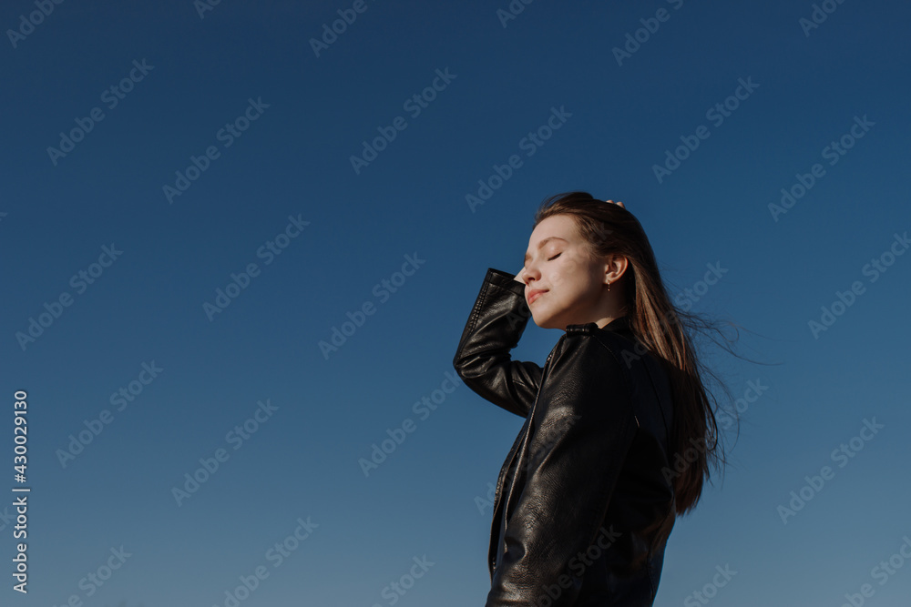 Smiling young woman in black jacket standing in front of blue sky enjoying bright sunlight touching hair. Mental health, relaxation, freedom. Girl in casual outfit with closed eyes. Golden hour sun Smiling young woman in black jacket standing in front of blue sky enjoying bright sunlight touching hair. Mental health, relaxation, freedom. Girl in casual outfit with closed eyes. Golden hour sun