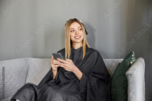 Happy beautiful light hair caucasian woman at beauty salon is sitting on sofa and using smartphone