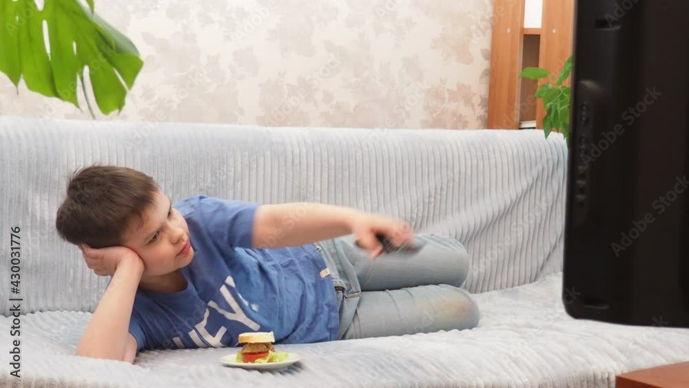 a teenage boy at home lies on the couch and watches TV, cartoons or movies, switches channels, programs. Eating a hamburger. Free time. Leisure. modern youth a teenage boy at home lies on the couch and watches TV, cartoons or movies, switches channels, programs. Eating a hamburger. Free time. Leisure. modern youth