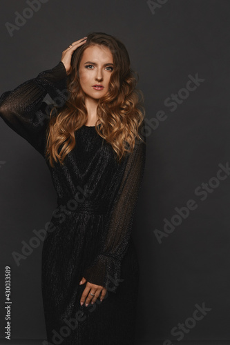 A model girl with blond curly hair and perfect makeup wearing a long evening dress posing over black background