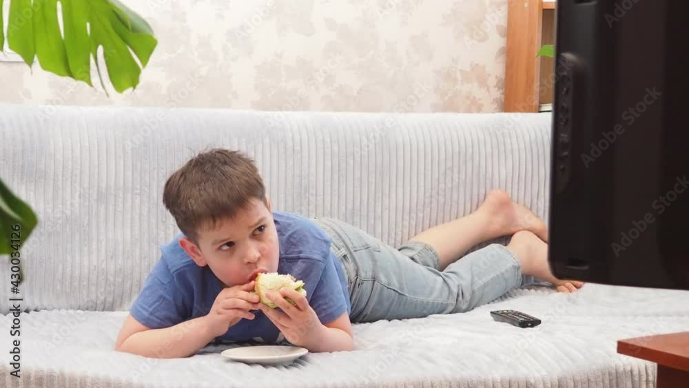 a teenage boy at home lies on the couch and watches TV, cartoons or movies, switches channels, programs. Eating a hamburger. Free time. Leisure. modern youth a teenage boy at home lies on the couch and watches TV, cartoons or movies, switches channels, programs. Eating a hamburger. Free time. Leisure. modern youth