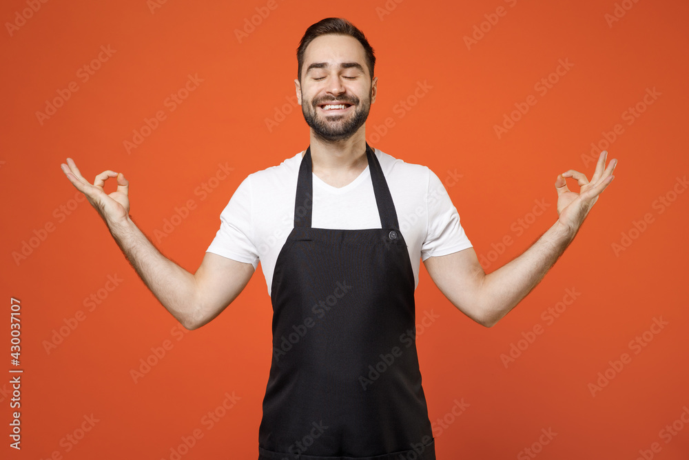Fototapeta premium Young man 20s barista bartender barman employee wear black apron white t-shirt work in coffee shop spread hands in yoga om gesture relax meditate isolated on orange background Small business startup.
