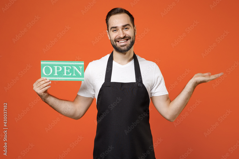 Young man barista bartender barman employee in apron white t-shirt work in coffee shop point index finger on card sign OPEN title text spread hand isolated on orange background Small business startup. Young man barista bartender barman employee in apron white t-shirt work in coffee shop point index finger on card sign OPEN title text spread hand isolated on orange background Small business startup.