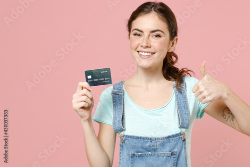 Young positive happy caucasian woman 20s in trendy denim clothes blue t-shirt pointing index finger on credit bank card show thumb up gesture isolated on pastel pink color background studio portrait