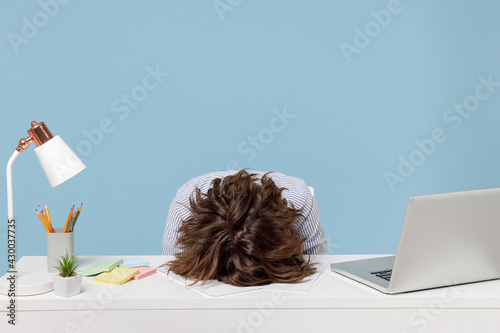 Young tired exhausted frustrated secretary employee business woman wearing casual shirt sit work sleep laid her head down on white office desk with pc laptop isolated on pastel blue background studio.