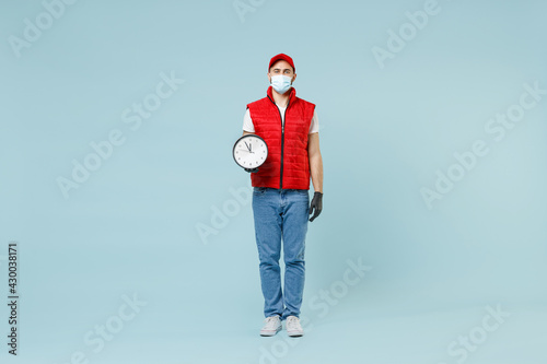 Full length delivery guy employee man in red cap white t-shirt vest uniform sterile face mask gloves work courier service on lockdown covid-19 flu virus hold clock isolated on pastel blue background.