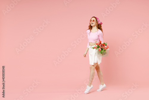 Full length young smiling happy stylish caucasian woman 20s in rose clothes bandana glasses hold tulips flowers bouquet walk isolated on pastel pink background studio portrait. Spring season concept.